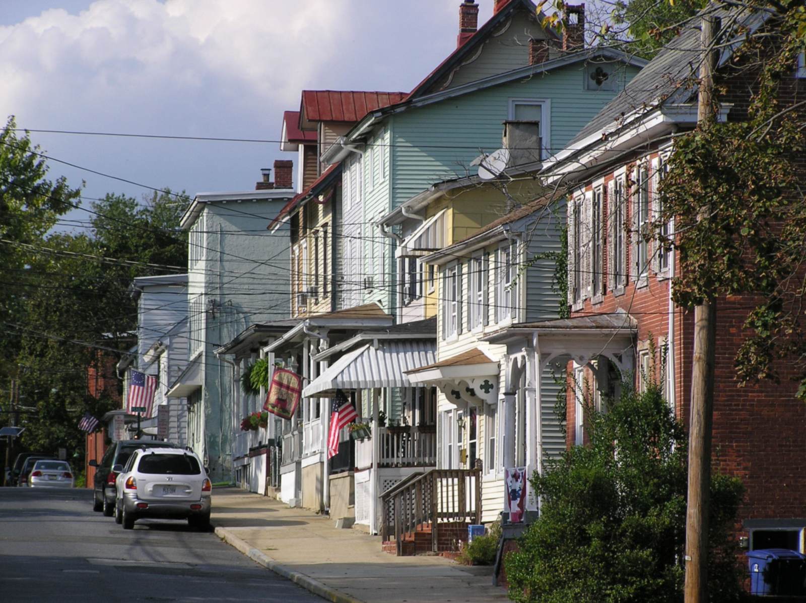 Row of attached two- and three-story houses in pastel siding and red brick, with front porches, striped metal awnings and American flags, along a narrow street with cars parked at the curb