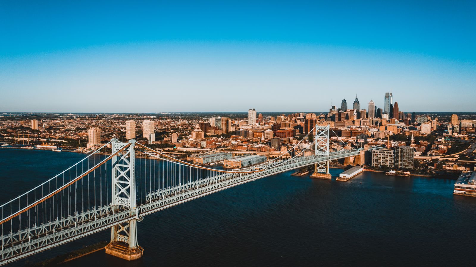 Aerial view of the Ben Franklin Bridge crossing the Delaware River toward the Philadelphia skyline under a clear blue sky