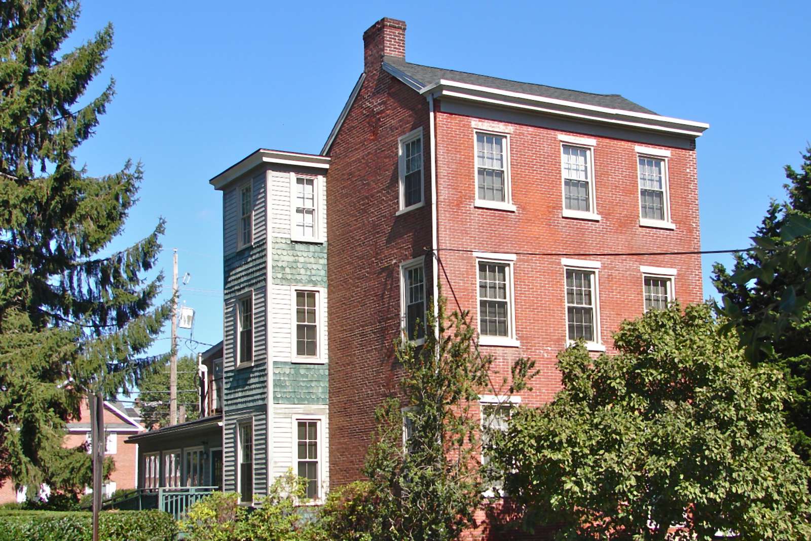Three-story red brick house with white-trimmed windows and a narrow clapboard wing with green fish-scale shingles, behind a spruce tree and leafy shrubs under a clear blue sky