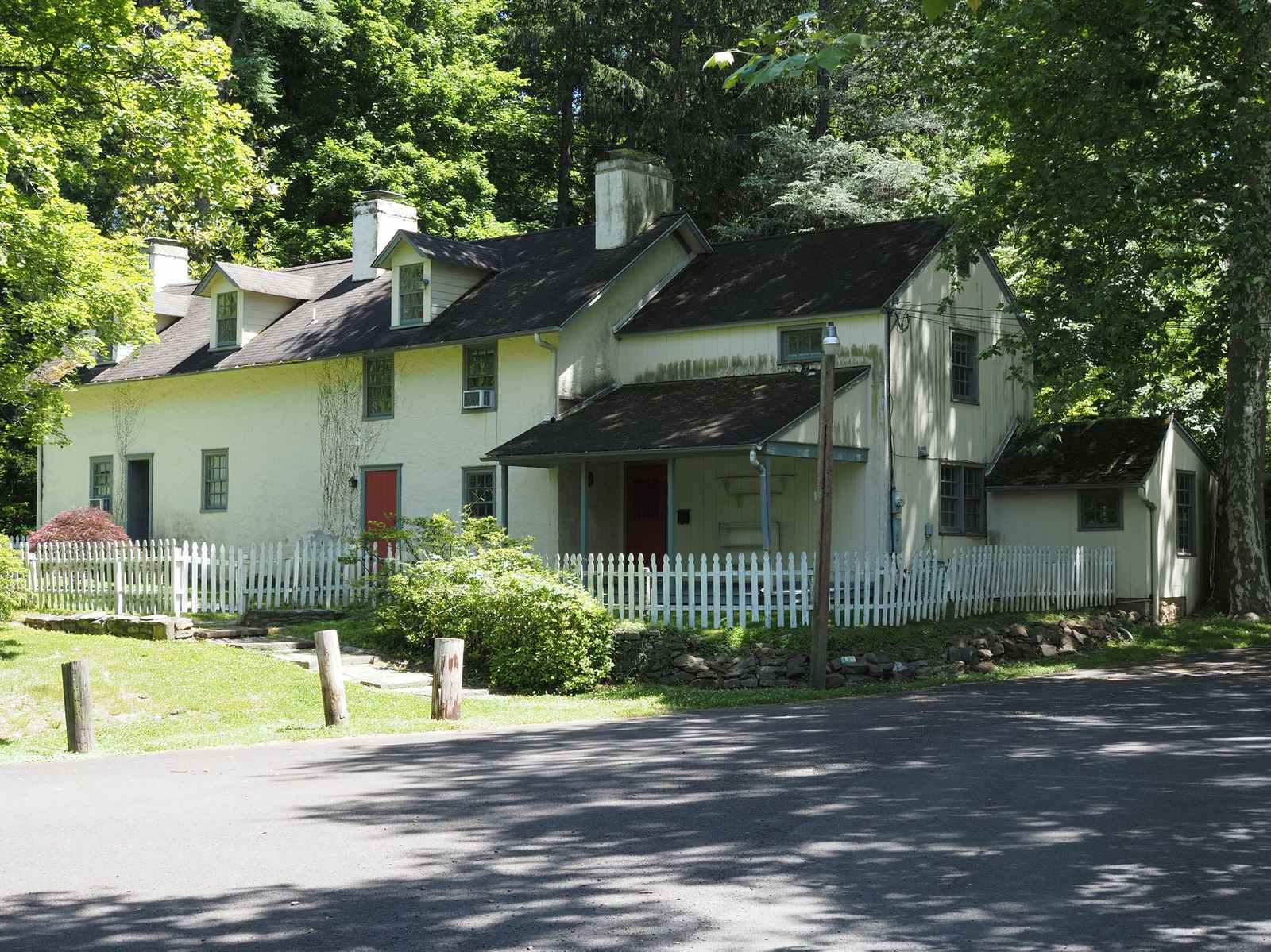Two-story cream-colored stucco house with dormers, red doors and a white picket fence, shaded by tall trees beside a paved lane