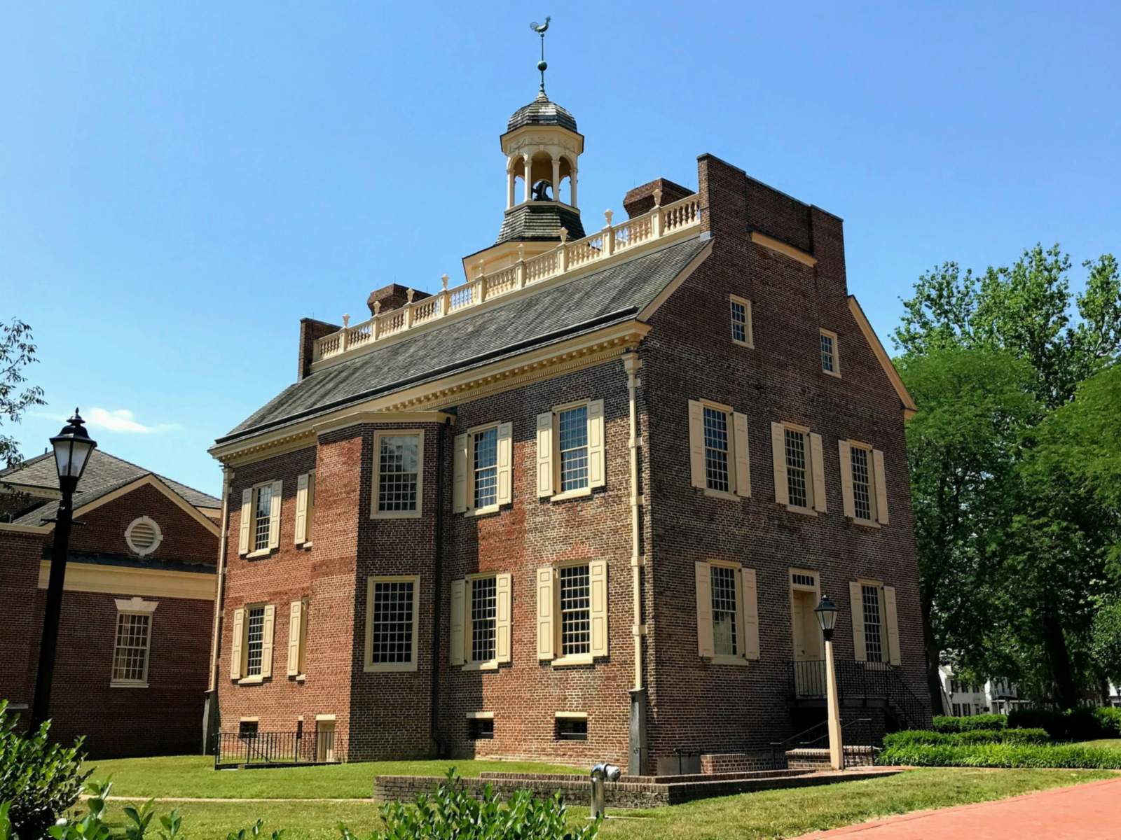 Three-story red brick colonial building with cream shutters, a balustraded roof and a domed cupola, beside a lawn and brick path under a clear blue sky