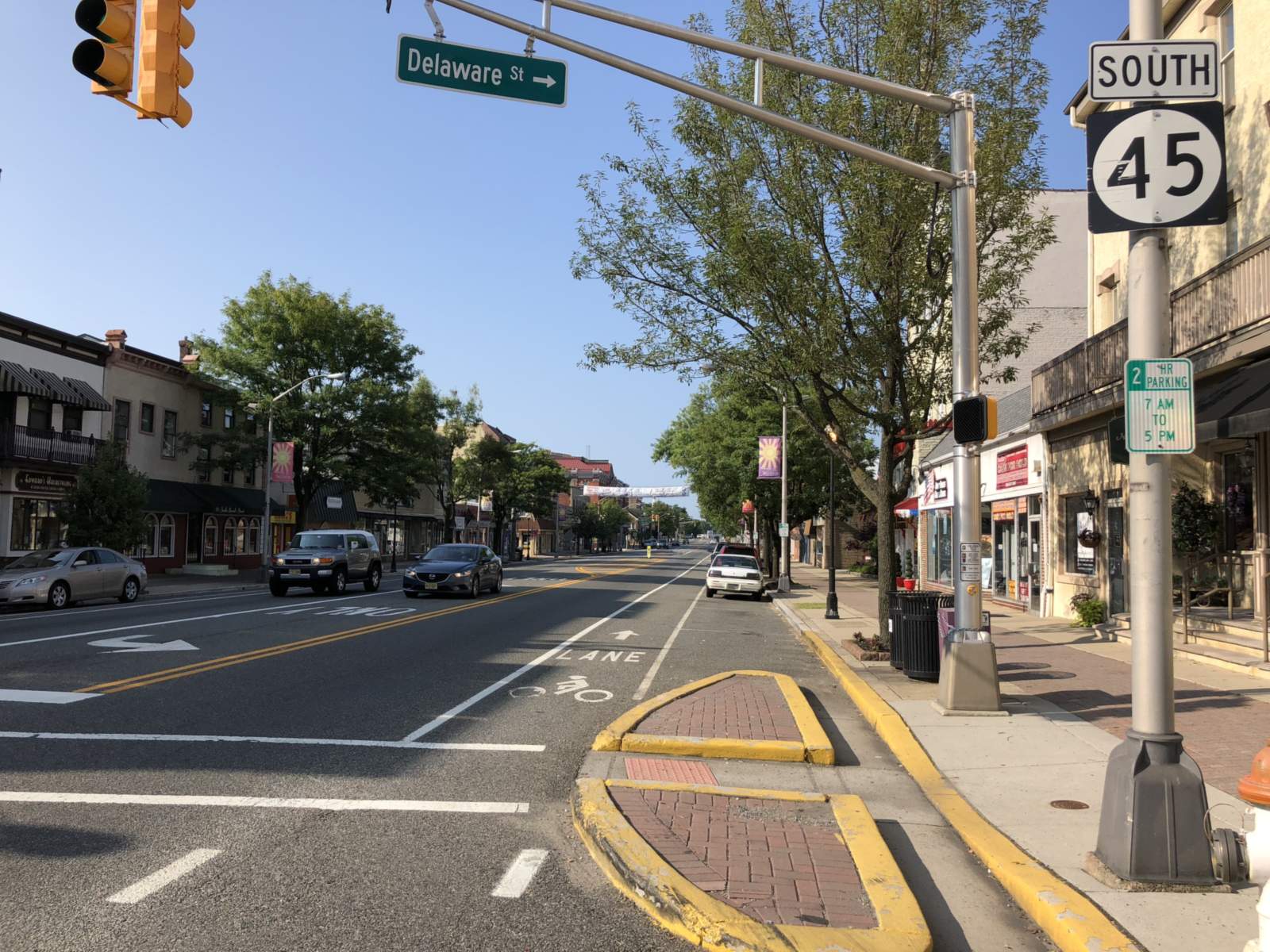 Main street signed Route 45 South at the Delaware Street corner, with a bike lane, brick-paved curb islands, and two- and three-story shopfronts under street trees on a clear morning