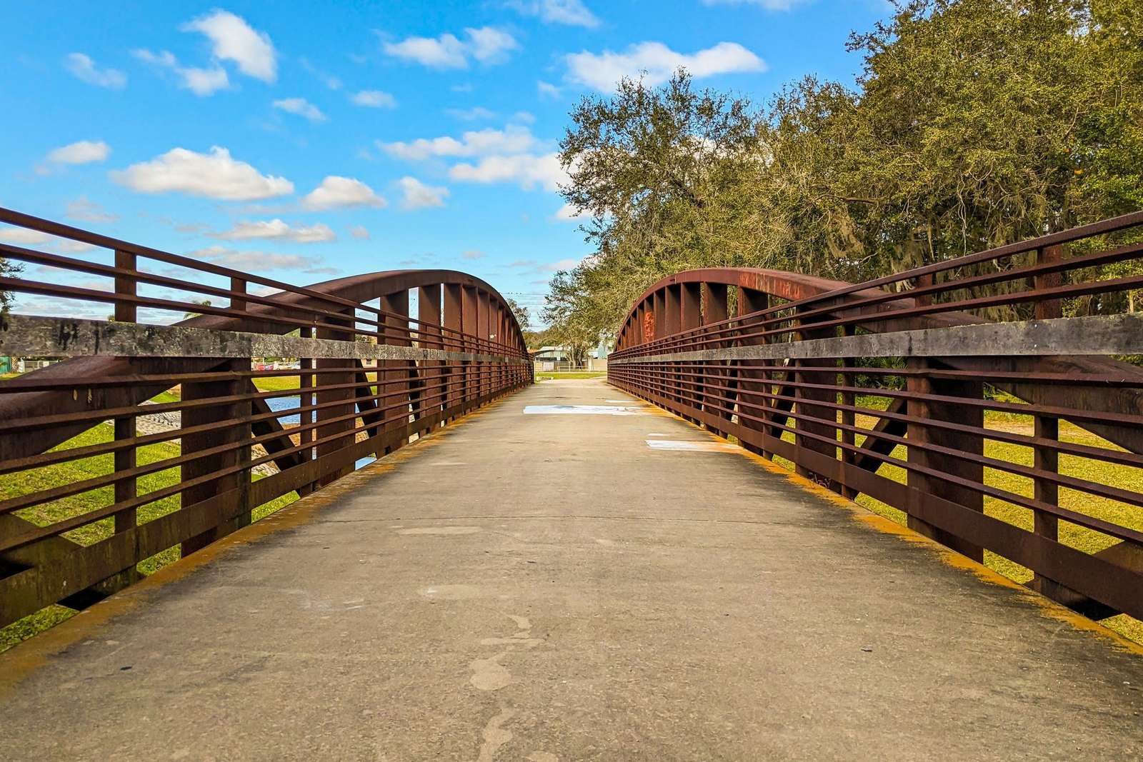 Concrete path crossing a rust-brown steel arch footbridge in a Kissimmee park, with grass, live oaks and a blue sky with scattered clouds
