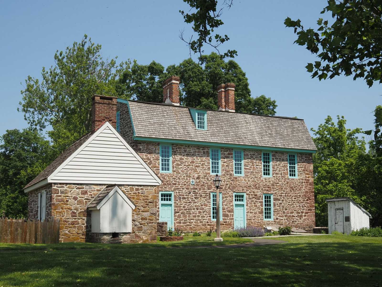 Two-story fieldstone house with turquoise window trim and a wood-shingle roof, a white-sided wing and a small white shed on a mowed lawn under tall trees