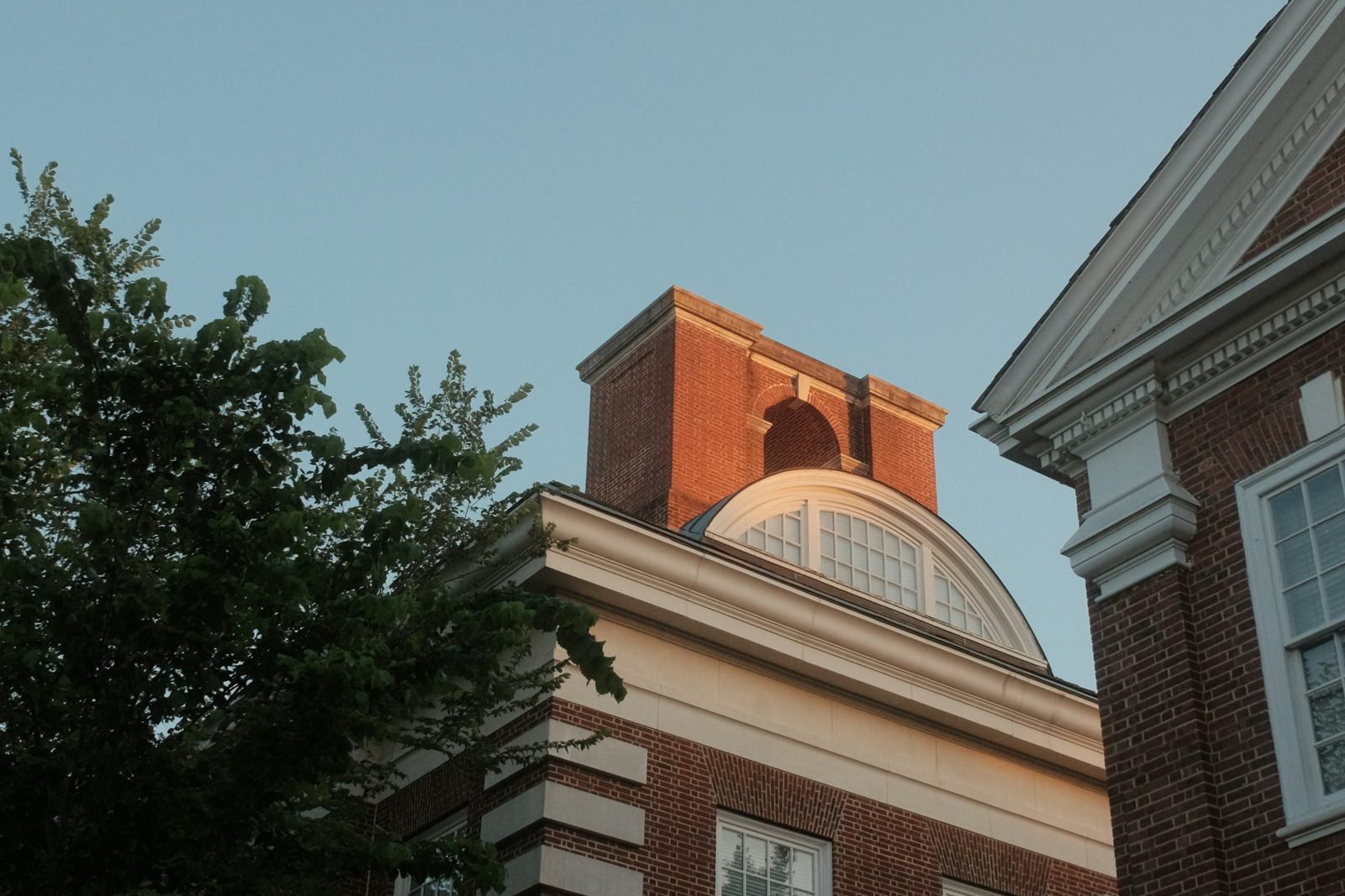 Upper floors of a red brick building with an arched white dormer and a brick cupola, lit by evening sun, with a leafy tree at left and a neighboring brick building at right