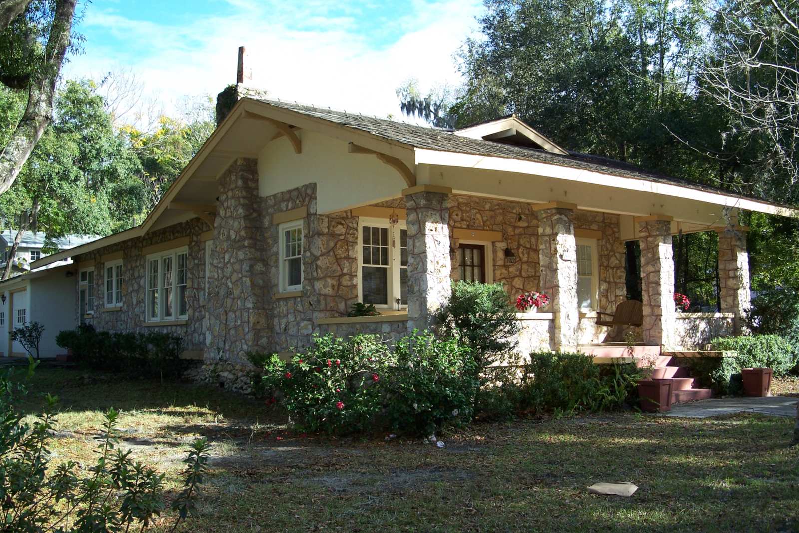 Single-story bungalow with walls of rough cream-colored stone, square stone porch columns, a wide low-pitched shingle roof and flowering shrubs in a shaded front yard