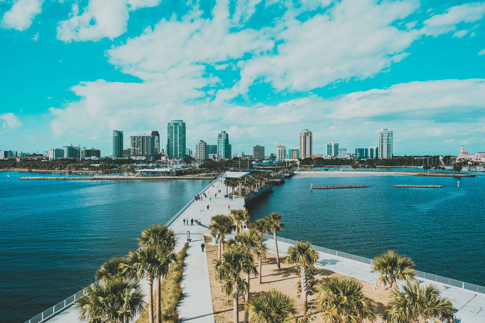 View from the St. Pete Pier back toward the downtown St. Petersburg skyline, with a palm-lined walkway, people strolling and blue water on both sides