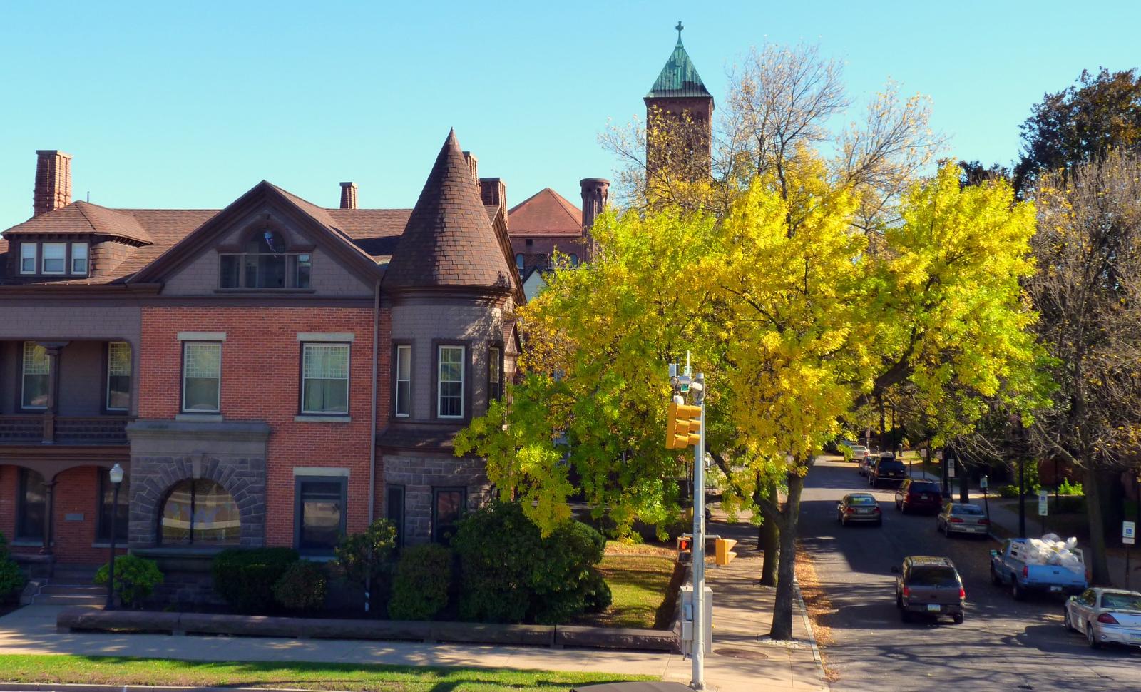 Brick and shingle Victorian house with a pointed corner turret on a tree-lined street, a yellow-leaved tree and parked cars beside it, and a church bell tower behind