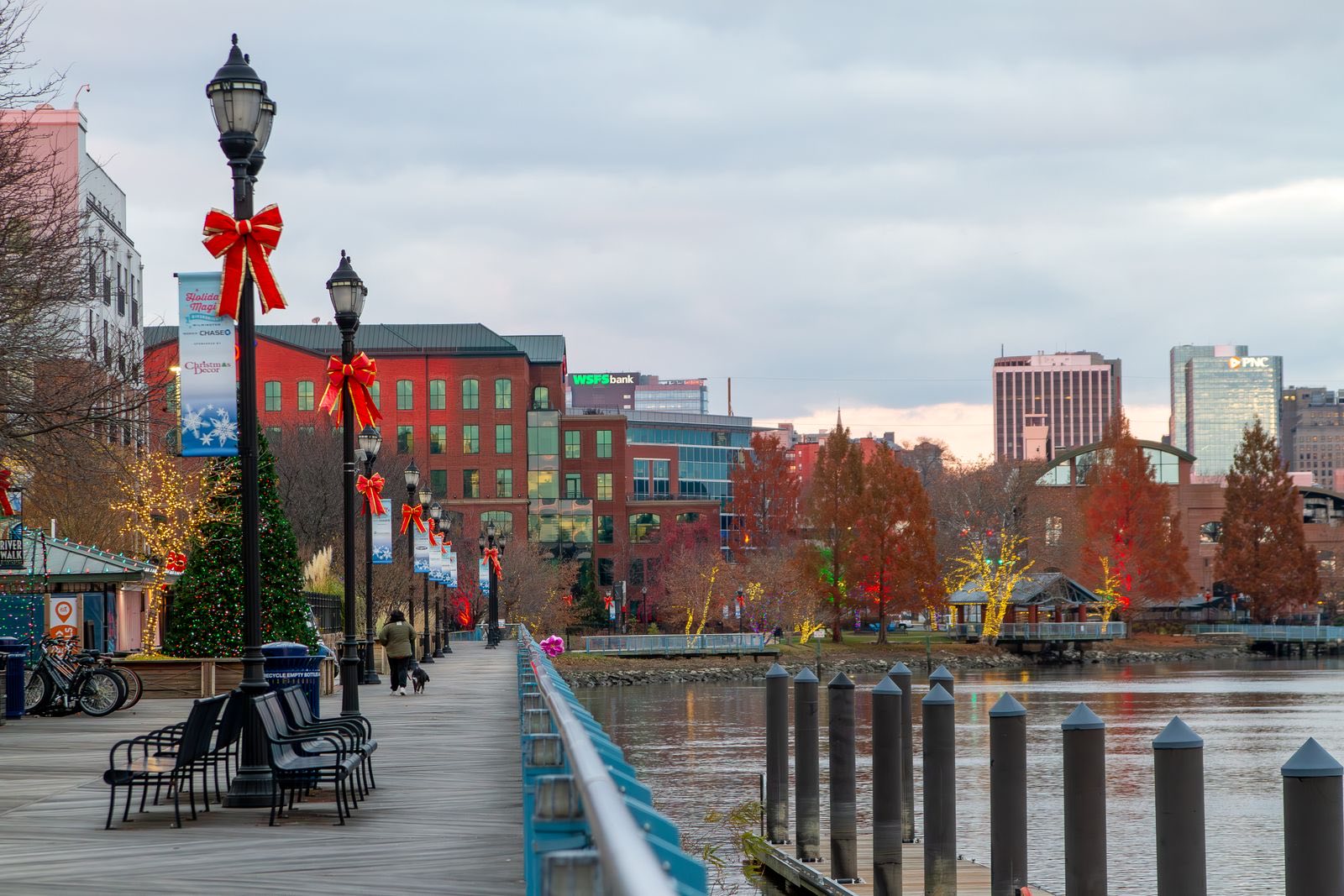Riverfront boardwalk in Wilmington with lamp posts tied with red bows, a decorated tree and office buildings beyond the river