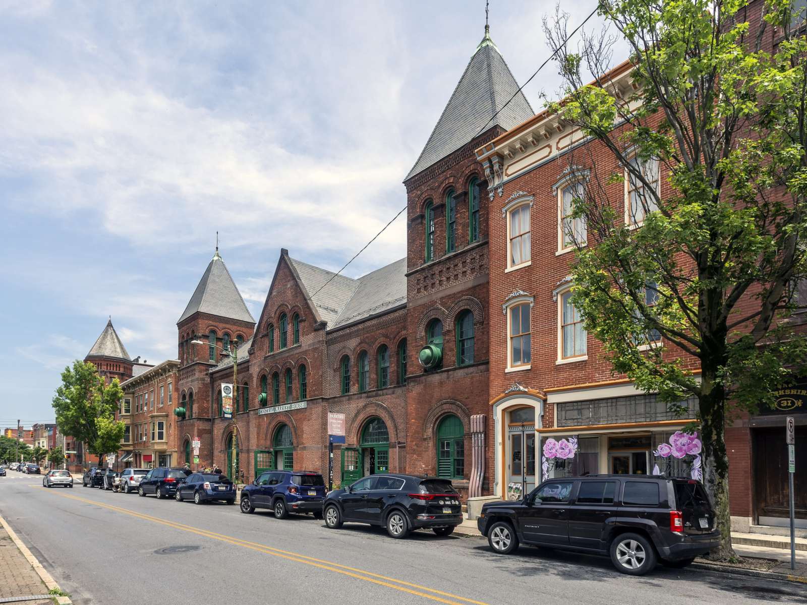 Red-brick market building with slate-roofed towers and green-trimmed arched windows beside brick rowhouses, with cars parked along a two-lane street
